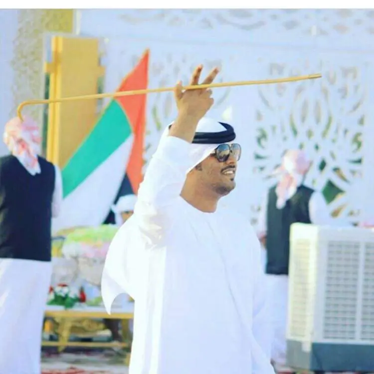 Emirati man in a white kandura raising a thin cane while dancing the ayala at a celebration with the UAE flag behind